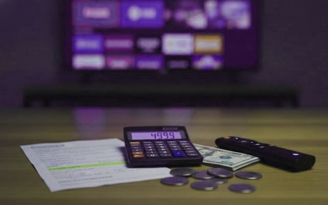 A calculator, dollar bills, coins, a streaming stick and a remote on a table in front of a TV